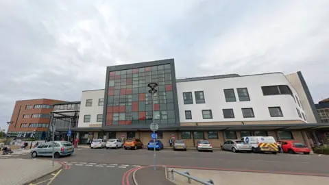 A modern hospital building, including a central glass fronted rectangular frontage next to white spaces on either side