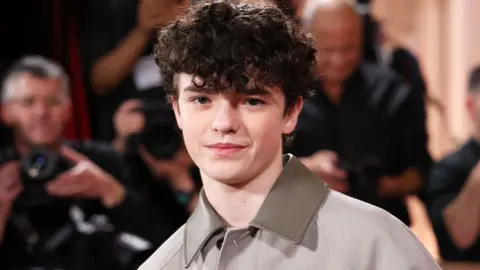 Owen cooper looking at the camera wearing a grey jacket with olive green leather collar. He is on the red carpet at the Golden Globe awards 