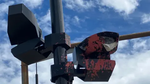 A close-up view of a roadside ULEZ enforcement camera mounted on a pole. Red paint has been splashed across part of the camera housing and support fittings. Blue sky and clouds can be seen in the background.