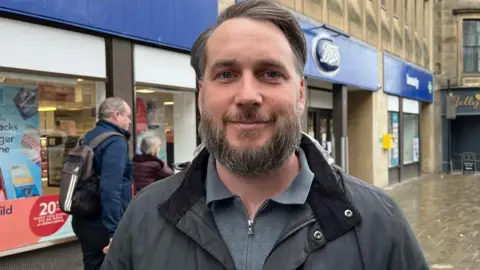 Grant Falconer has dark hair and a beard. He is wearing a grey outdoor jacket over a grey top and is standing in a street with shops behind him.