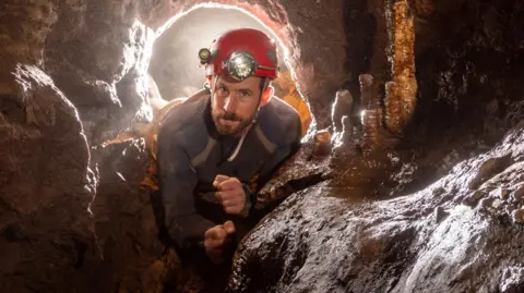 John Bolton John Bolton, a young man with a brown beard and blue eyes is wearing a red helmet with a head torch. He is in a cave, with stalagmites to his side.