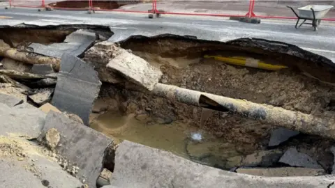 Adrian Harms/BBC The inside of the sinkhole, as viewed from surface level. The ground beneath the road surface is visible and water flowing out of a hole in a broken pipe is flowing into a pool at the bottom of the sinkhole.