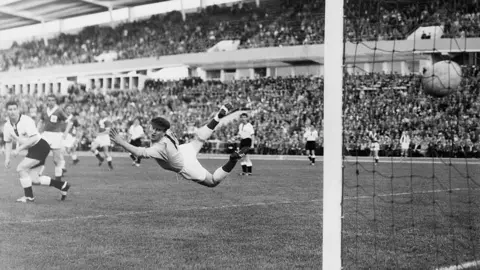 Getty Images A black and white photo from the 1958 World Cup showing Northern Ireland's goalkeeper Harry Gregg wearing a cap but no gloves diving to try to stop a shot from a German player who is not in shot. He is diving in mid air. The ball has flown past him and can be seen hitting the back of the net at the far right of the image. The game is being played in a stadium packed full of fans.
