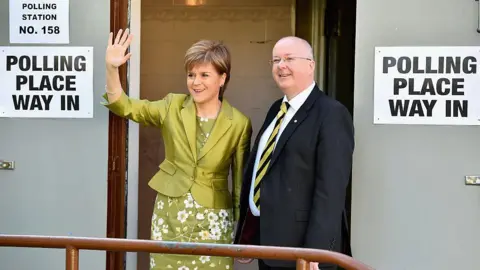 Getty Images Nicola Sturgeon in a green floral dress and matching cropped jacket. She is standing next to Peter Murrell, dressed in a dark suit, white shirt and black and yellow striped tie. They are standing outside a polling place on election day, posing for the cameras.