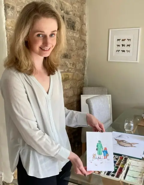 Eleanor Tomlinson smiling at the camera, holding the viral piece of art. She has shoulder length blonde hair and is wearing a white blouse and black trousers. 