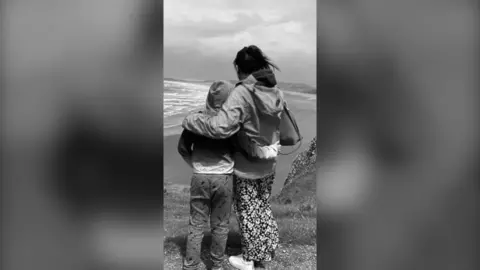 A woman hugs a boy as they look out of a beach