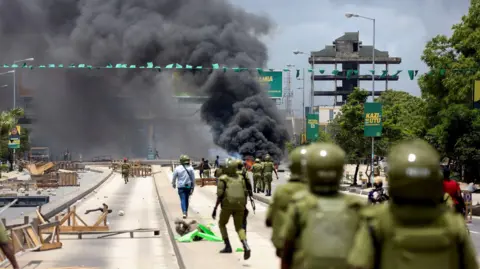 Reuters Armed police officers walk down a road strewn with debris. Flames and thick black smoke can be seen in the background.