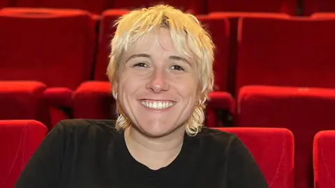 Maisie has short, shaggy blond hair and sits smiling in a theatre auditorium, leaning forward with hands clasped. She is wearing a black T‑shirt and sits among rows of bright red seats that fill the background.