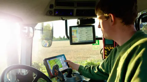 Getty Images A farmer in a tractor, using several digital devices including a GPS and a tablet.