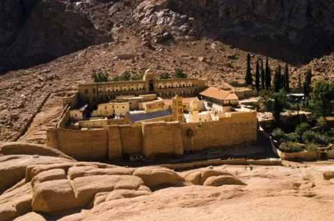 Universal Images Group via Getty Images The St Catherine's Monastery as seen from the outside. It is a walled structure with several buildings inside, with the monastery's gardens attached to one side, which are green with some trees inside. The surrounding area is rocky with the ground sloping up behind the monastery at the foot of a mountain 