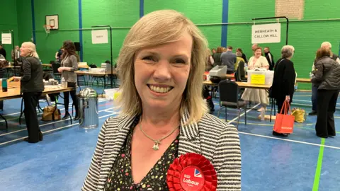A woman smiling at the camera, wearing a red Labour rosette.