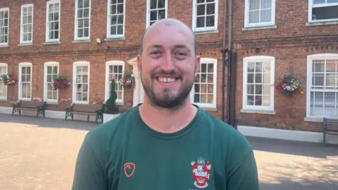 A man with a shaved head and a beard, wearing a green t-shirt with a crest on from RGS Worcester stands in front of a large brick building with large white frame windows.