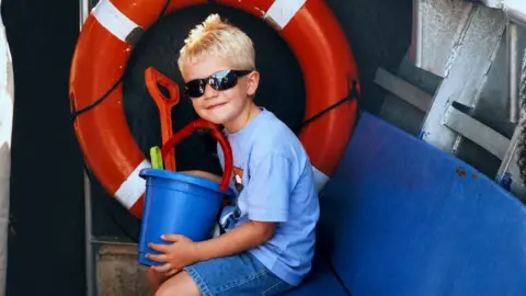 A childhood photograph showing a young Max Golding sitting inside the Mudeford Ferry holding a bucket and spade. An orange life ring hangs on the wall behind.