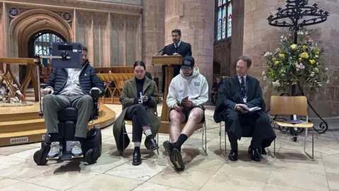 The King's School Ed Slater sitting in his motorized wheelchair on the left. He is joined by his wife Jo sitting on a wooden chair beside him, and Lewis Ludlow sitting beside her. They are inside a cathedral near the pulpit while someone delivers the assembly behind them. There is a big display of flowers on the right.