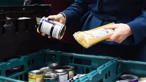 A woman holds a packet of spaghetti in her left hand and a tin in the right, above a stack of plastic food baskets.