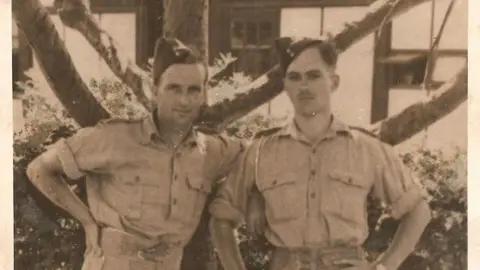 Woods Family Joe Woods and a fellow solider in military uniform standing leant on a tree.