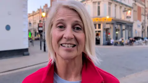 A woman with short, blonde hair standing in the street, smiling at the camera. She is wearing a red jacket. 