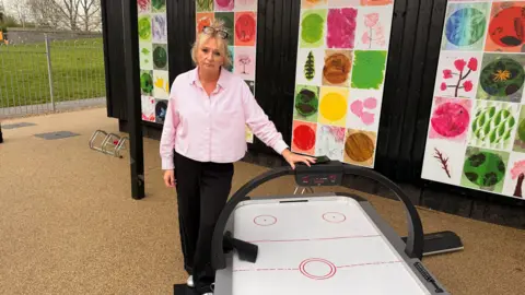 Jo Ramsay - a woman with blonde hair and glasses on her head. She is wearing a pink shirt and holding an ice hockey table that is broken. The table has collapsed and the woman looks upset about it. 