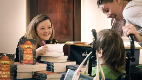 TVCA Author Lisette Auton at a book signing. She sits behind a desk covered in books while a child and parent wait for a signed copy of one of her books.