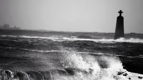 A stone cross on a plinth poking out from a wild sea.