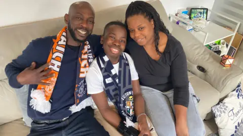 Kayden sits on the sofa with his mum and dad. Kayden is wearing a Luton Town scarf around his neck.