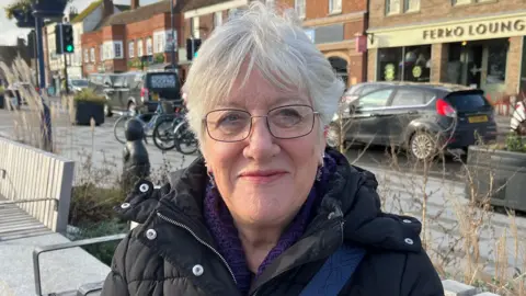 Tom Jackson/BBC Ruth Noonan is sitting on a bench. She has short grey hair and is wearing black frames glasses, a purple jumper and black coat. Behind her are cars queuing at lights. 