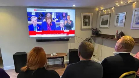 Shaun Whitmore/BBC Jenny Wilson, Nick Anema and Marvin Shalders pictured from behind, as they watch a large screen TV which should Rachel Reeves delivering her Budget.