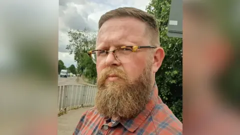 Liam Calvert Liam Calvert, wearing glasses and a beard, looks at the camera while standing on the street. He is wearing a red, orange and blue checkered shirt.