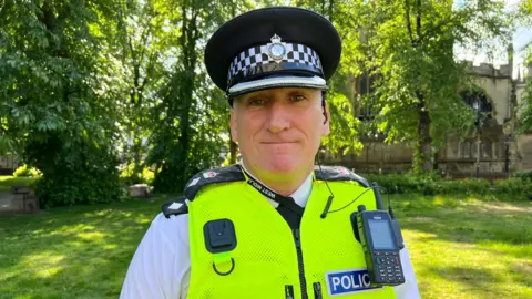 Ch Supt Paul Joyce looks at the camera wearing a flat police cap, yellow high visibility vest with a walkie talkie on the left side of his chest and the word police in white writing with a blue background beneath. He is standing outside a church surrounded by greenery.