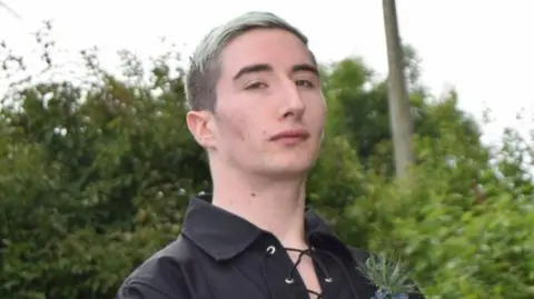 Spindrift Thomas Fraser has blonde dyed hair swept to one side. He is wearing black dress shirt usually worn with a kilt.