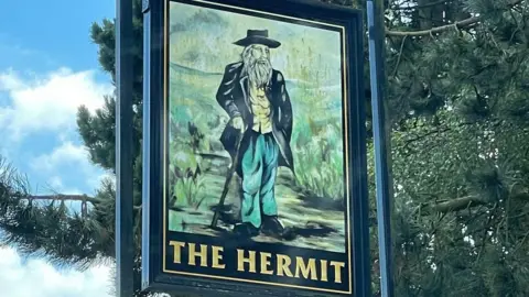 The Hermit Inn A pub sign showing what is obviously meant to represent an eccentric old man with flowing beard, walking cane and colourful though scruffy attire