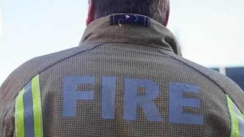 PA Medium A shot of a firefighter taken from behind his shoulders. On the top of his light brown jacket it reads: "Fire". He is looking up at a gray sky.