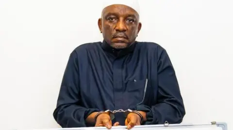 Saleh Mamman wearing a black cloth, handcuffed and holding a white board
