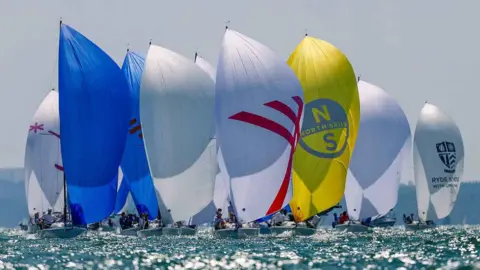 A fleet of racing yachts sail with colourful sails raised during competition on the Solent.
