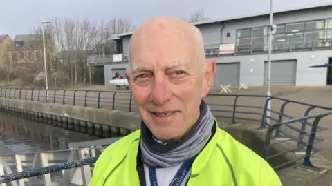 Ralph Pickles is wearing a yellow jacket, white top, grey and black scarf and is stood next to water. There is a grey boathouse behind him.