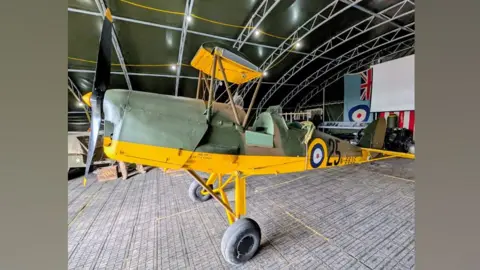 Bottisham Airfield Museum Tiger Moth N6635 in a hangar. It has a grey body with a bright yellow underbelly but its two wings are missing.