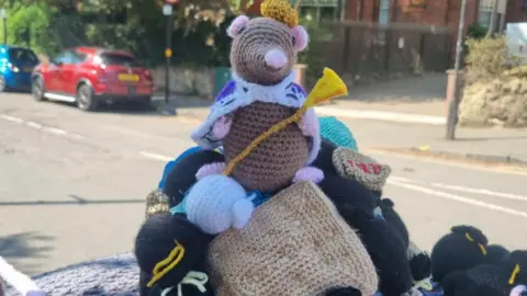 Moseley Knit and Natter Group A knitted rat, with a crown and a sceptre designed to look like cheese, sits on top of a mound of knitted bin bags on top of a postbox.