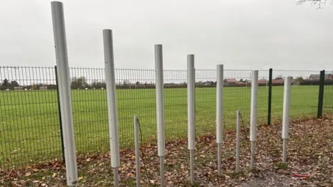 Luke Deal/BBC The image shows an outdoor musical installation consisting of seven tall, cylindrical metal tubes arranged in a row along a dirt path. 