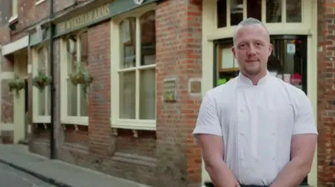 Chef Luke Emmess standing outside the Wykeham Arms pub in Winchester - it is a brick building on a corner. Luke is wearing chef's whites.