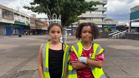 A young girl with long brown hair tied back stands in a black tshirt and hi-vs jacket. A young boy with brown curly hair and a braid in it stands with his arms crossed and a red football shirt on and a hi-vis jacket on top. Behind them is a paved square and shops with high rise flats behind.