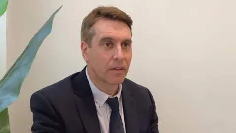 Giles Emmanuel is in a minimally decorated room with a green plant beside him. He has short brown hair and is dressed in a dark blazer, a light shirt and a navy tie.