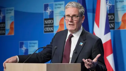 Keir Starmer gestures with his hand while speaking at a Nato summit on Wednesday in the Hague, he's wearing a suit and tie and glasses and stands at a podium with the UK flag in the background against a blue wall with the Nato logo on.