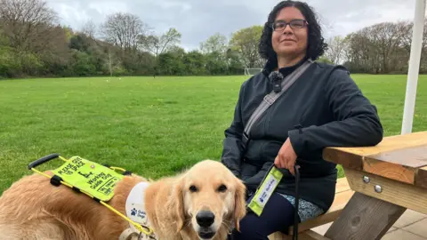 A woman in a black jacket sitting on a picnic bench in front of a field with a golden Labrador standing in front of her. The dog is wearing a harness describing it as a a working guide dog.