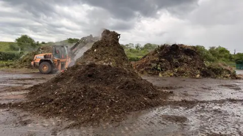Brian Seeger A large pile of compost in a muddy yard. There is a digger in the background next to another pile of large compost. It is a cloudy day. There are fields and trees in the background.