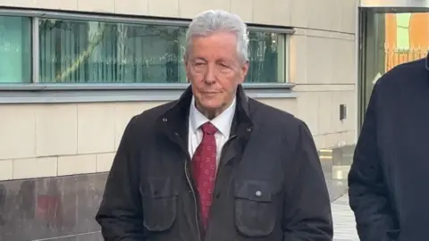 BBC Peter Robinson standing in front of the court. He has grey hair and is wearing a shirt and red tie under a black coat.