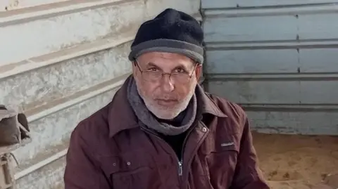 Khalid El-Astal A man is pictured sitting in a metal structure. He has a grey beard and is wearing glasses, a dark-coloured hat , a maroon coat and dark top. 