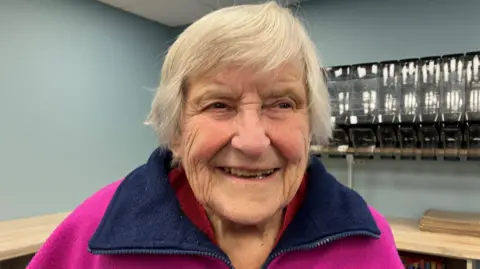 An older lady with white hair smiling. Wearing a purple fleeced with a blue neck. 