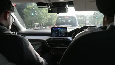 Two police officers with their backs to the camera sit in the front two seats of a police car as it drives through central London.