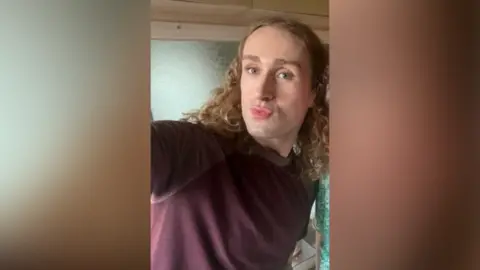 West Mercia Police A person with shoulder-length dark blonde curly hair pouts while taking a selfie while wearing a burgundy jumper.