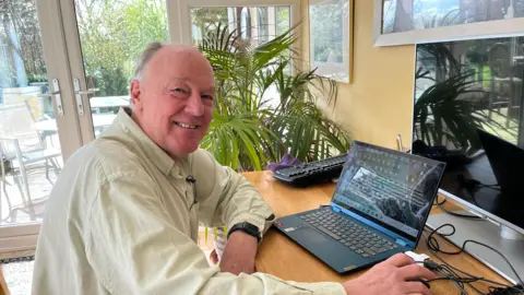 Martin Surl, wearing an off-white shirt and smiling at the camera. He is sitting in front of a laptop as if working , with his hand on the mouse. Behind him are glass patio doors with patio furniture in the garden. Behind Surl are some indoor plants.